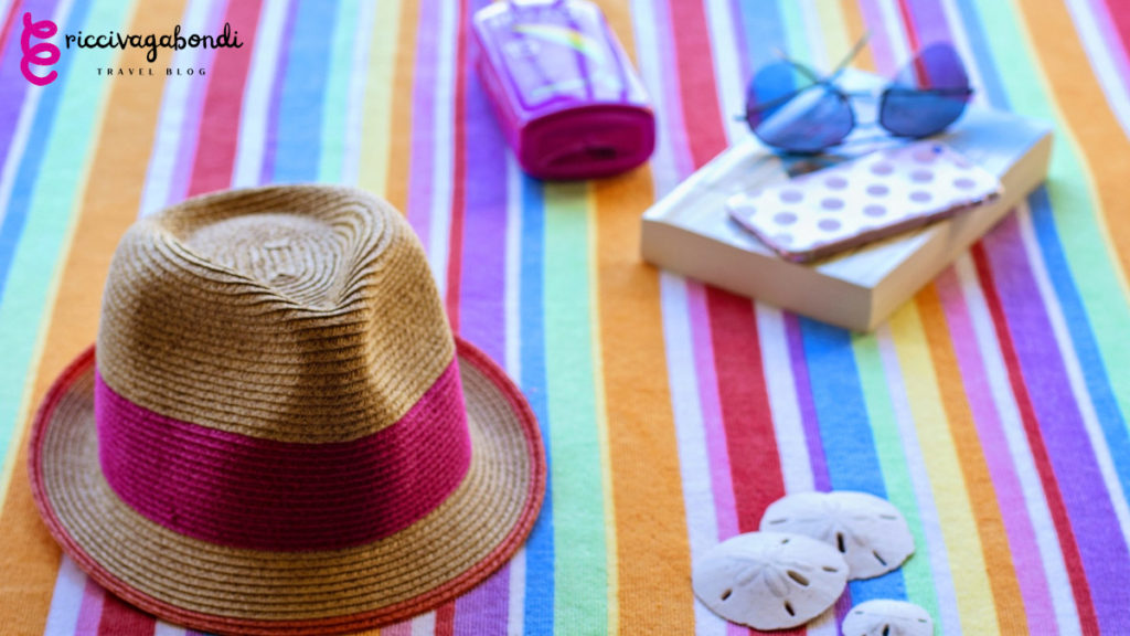 A hat, sunglasses, phone, sunscreen, book and some shells on a colourful plaid